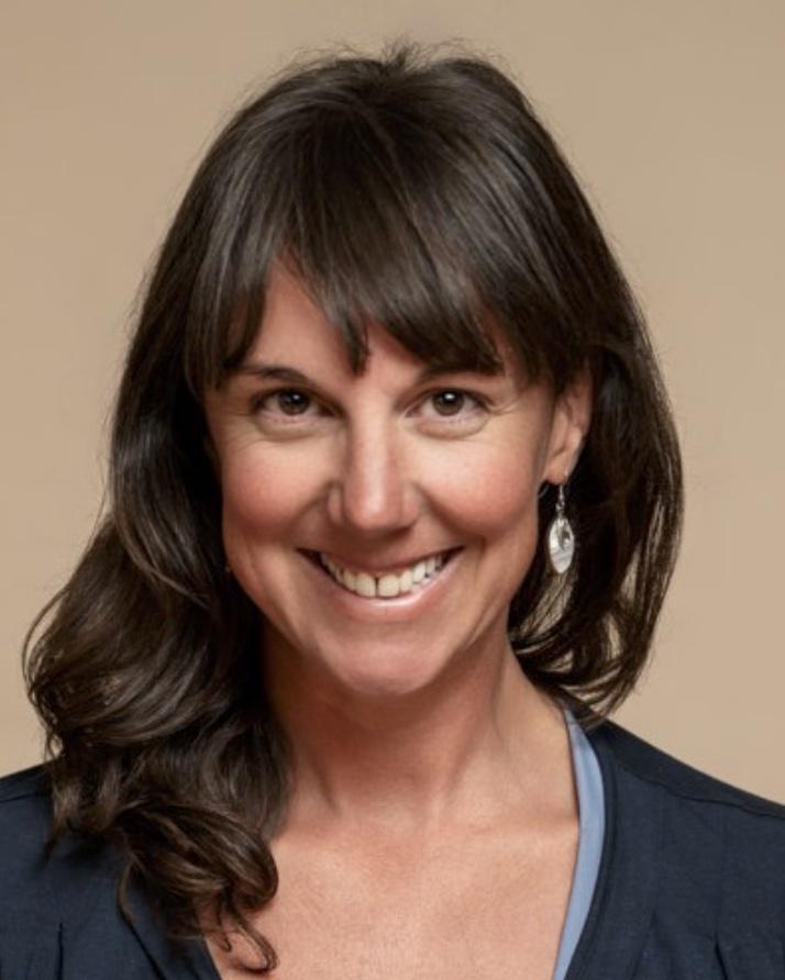 Portrait of Allison smiling, with dark hair and a navy top, against a warm beige background